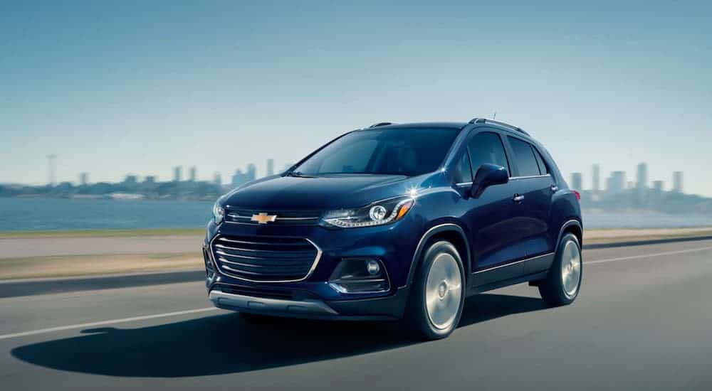 A blue 2019 Chevy Trax is driving past a body of water and a city skyline.