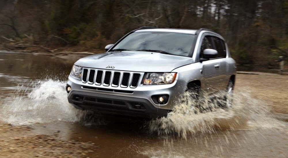 A silver 2013 Jeep Compass is driving through a puddle in the woods after leaving a used Jeep dealer.