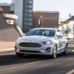 A white 2020 Ford Fusion is driving on a city street as part of the 2020 Ford Fusion vs 2021 Honda Accord comparison.