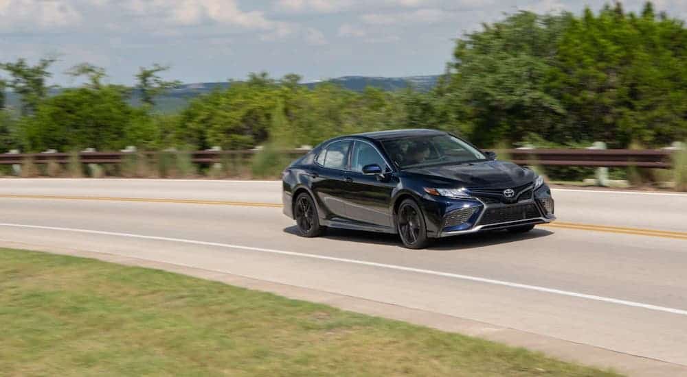 A black 2021 Toyota Camry is driving on a highway in front of trees.