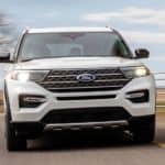 A white 2021 Ford Explorer King Ranch is shown from the front driving on a road in front of yellow grass and a tree.