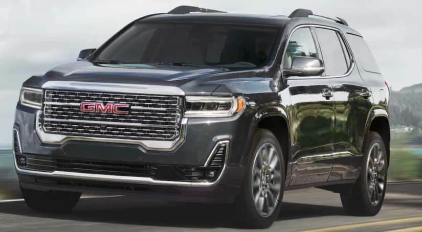 A dark grey 2021 GMC Acadia is driving past a body of water after winning the 2021 GMC Acadia vs 2021 Kia Telluride comparison.
