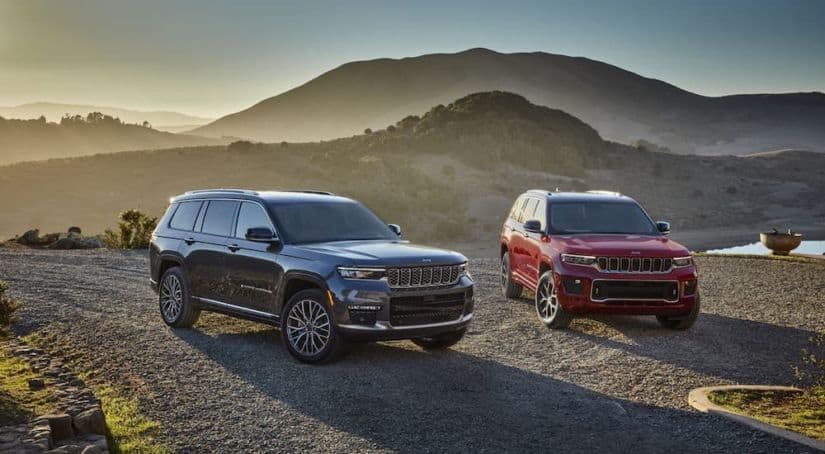 A red and a gray 2021 Jeep Grand Cherokee L is parked in front of distant mountains.