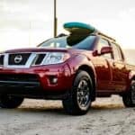 A red 2021 Nissan Frontier with a surfboard on the roof is driving away from a beach.