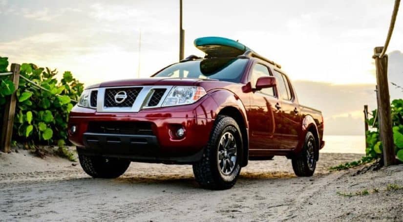 A red 2021 Nissan Frontier with a surfboard on the roof is driving away from a beach.