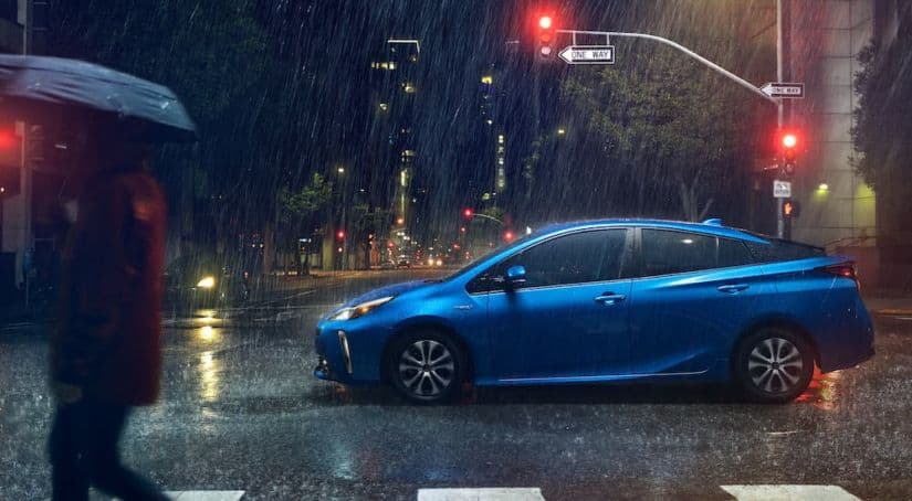 A blue 2021 Toyota Prius is shown from the side driving in a city in the rain at night.