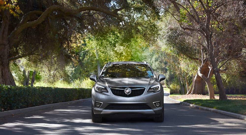 A silver 2021 Buick Envision is shown from the front driving down a tree-lined road.