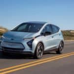 A pale blue 2022 Chevy Bolt EV is driving on a highway after leaving a local Chevy dealership.