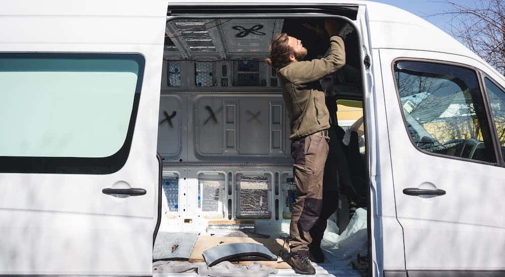 A man is shown inside the open door of a white commercial van that is being converted into a camper.