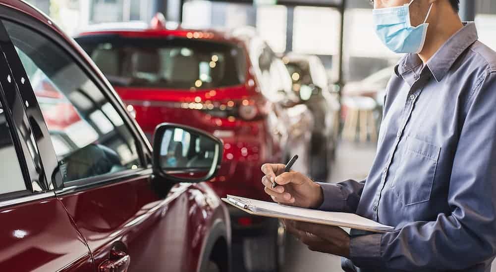 A salesman in a mask with a clipboard is raising prices on red vehicles in current auto news.