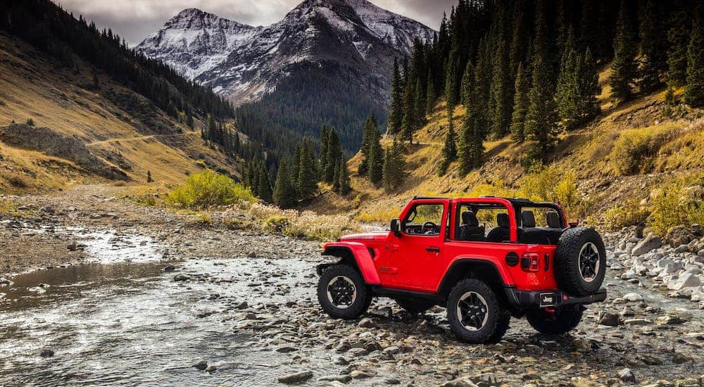 A red 2021 Jeep Wrangler with no roof is crossing a river in front of trees and distant mountains.