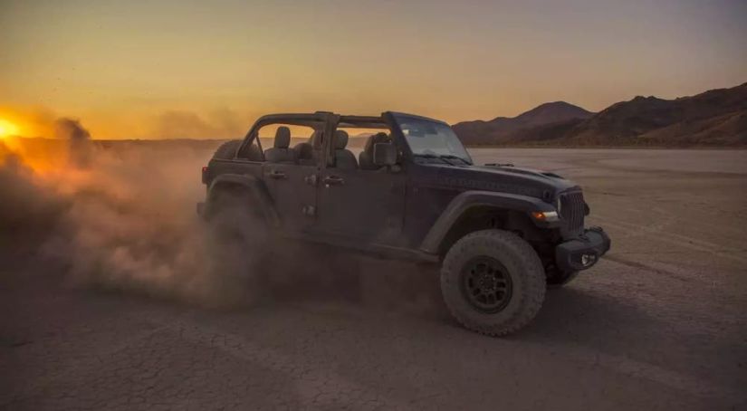 A black 2021 Jeep Wrangler Rubicon 392 with no roof is kicking up sand in a desert at sunset.