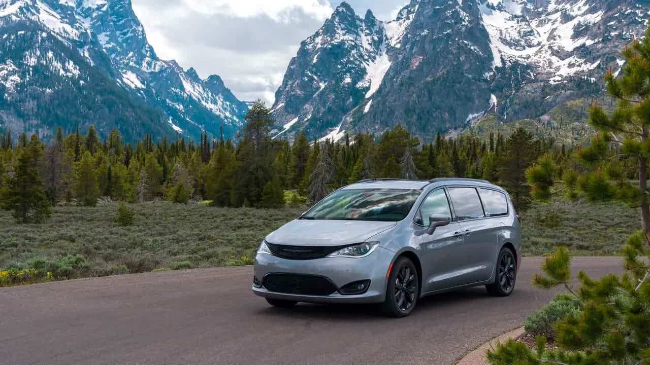 A silver 2018 Chrysler Pacifica is driving in front of evergreen trees and snow-covered mountains.