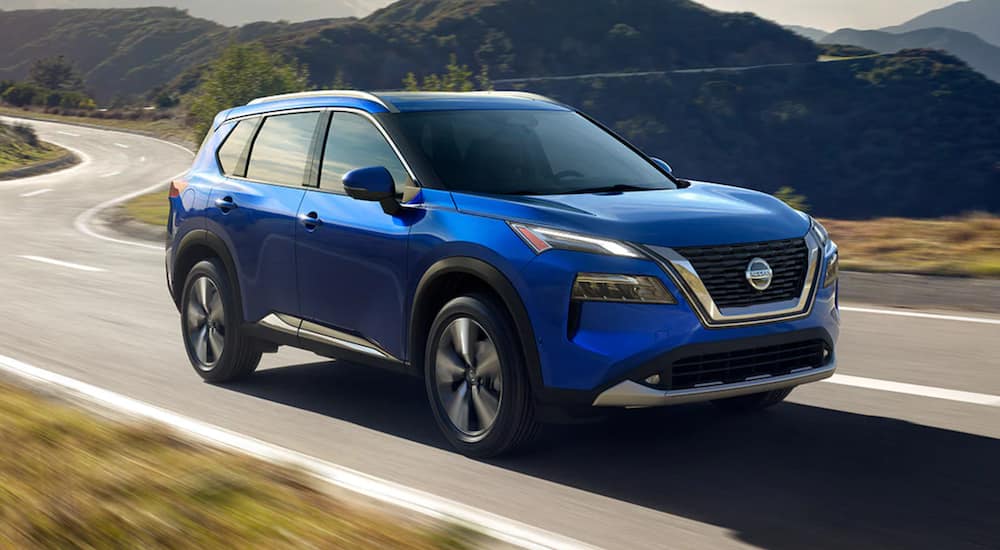 A blue 2021 Nissan Rogue is driving down an open road in front of mountains.