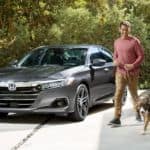 A man is walking his dog next to a grey 2021 Honda Accord in the driveway of a modern house.