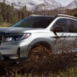 A silver 2021 Honda Ridgeline Sport has a dirt-bike in the bed while driving through a big mud puddle.