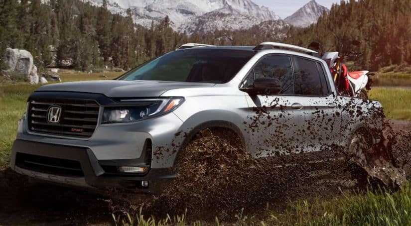 A silver 2021 Honda Ridgeline Sport has a dirt-bike in the bed while driving through a big mud puddle.