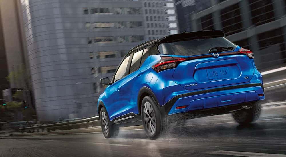 A blue 2021 Nissan Kicks is shown from the rear driving through a city on a wet road.