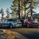 A light blue 2021 Toyota Highlander Platinum is next to a red Highlander hybrid parked outside of a cabin in the mountains.