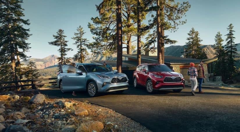 A light blue 2021 Toyota Highlander Platinum is next to a red Highlander hybrid parked outside of a cabin in the mountains.