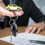 A salesperson in a suit is holding a yellow toy car and car keys over GMC lease paperwork,
