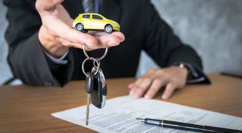 A salesperson in a suit is holding a yellow toy car and car keys over GMC lease paperwork,