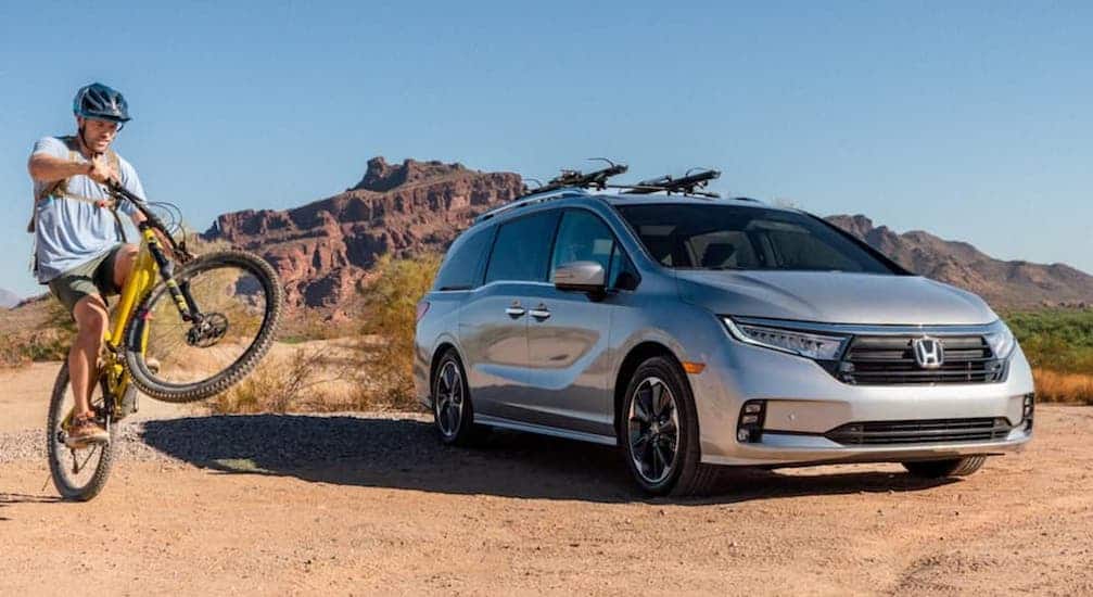 A silver 2021 Honda Odyssey is shown parked in the desert next to a man doing a wheelie on a mountain bike.