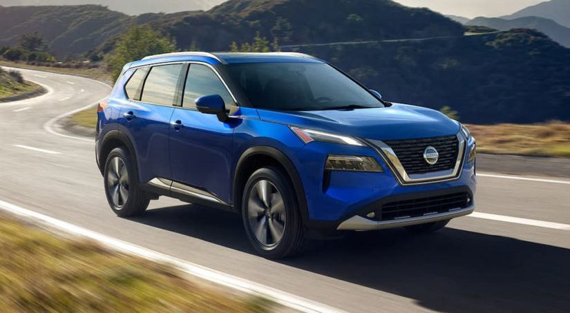 A blue 2021 Nissan Rogue is shown driving on a winding road in the mountains.