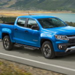 A light blue 2021 Chevy Colorado is driving on a two way road past a body of water after leaving a Chevy Dealer.