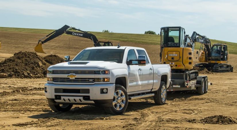 A white 2018 Chevrolet Silverado 2500HD is towing machinery in a sand pit.