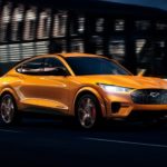 An orange 2021 Ford Mustang Mach-E is driving through a city at night after leaving a Ford Dealer.