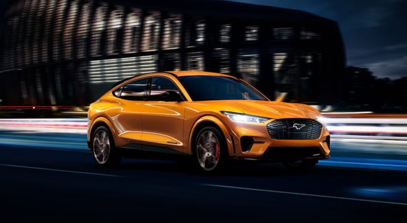 An orange 2021 Ford Mustang Mach-E is driving through a city at night after leaving a Ford Dealer.