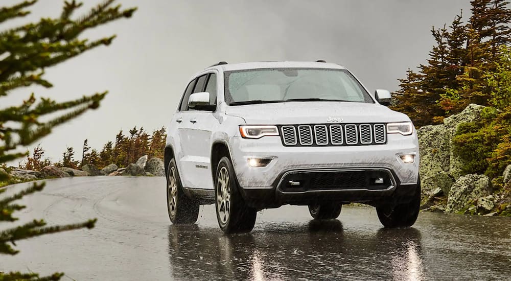 A white 2020 certified pre-owned Jeep Grand Cherokee is driving on a tree-lined road in the rain.