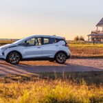 An upcoming Chevy EV, a pale blue 2022 Chevy Bolt EV, is shown parked on a brick path next to a beach house.