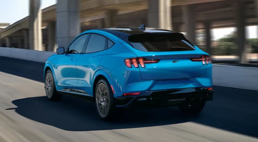 A blue 2021 Ford Mustang Mach-E is driving away from a NY Ford Mustang Mach-E dealer.