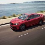 A red 2021 Subaru Legacy is driving next to the ocean after leaving a Subaru Legacy dealer.