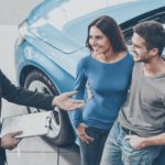 A couple is shown inside of a used SUV dealer talking to a salesman next to a car.