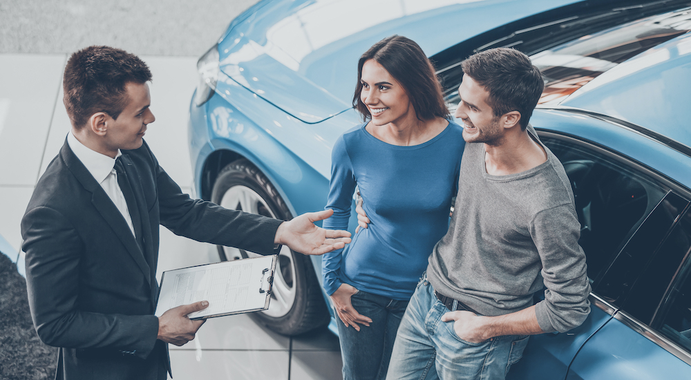 A high angle view of a used car salesman speaking to a couple leaning against a used car.