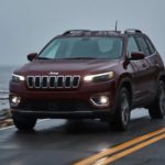 A used maroon 2020 Jeep Cherokee is driving next to the ocean after leaving a used SUV dealership.