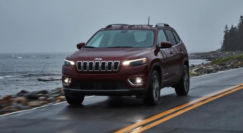 A used maroon 2020 Jeep Cherokee is driving next to the ocean after leaving a used SUV dealership.