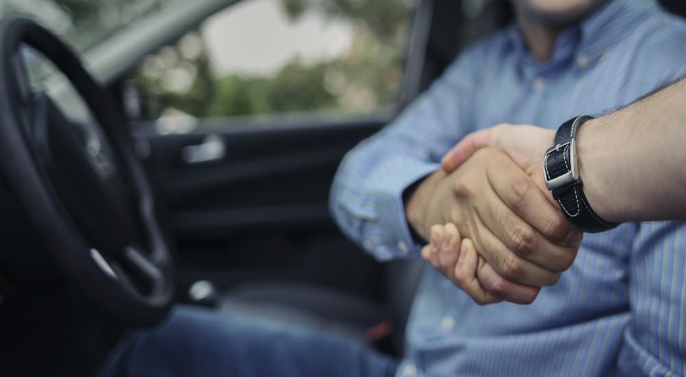 A man at a used car dealership is shaking hands with a salesman in the front seat of a car.