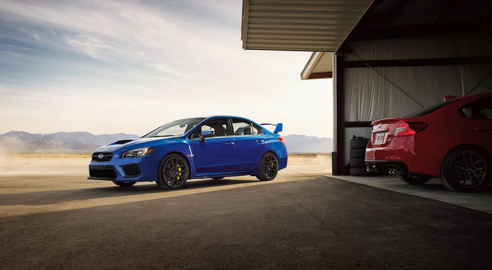 A blue 2021 Subaru STI is parked next to a red 2021 WRX in a shed.