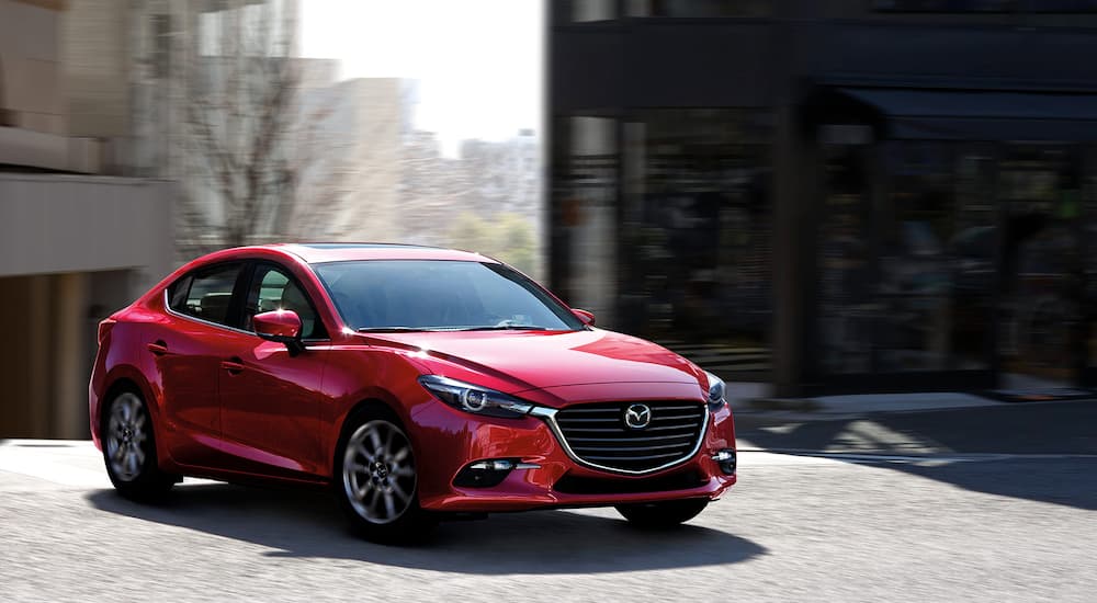 A red 2018 Mazda3 Sedan is shown driving past a shop after leaving a used car dealer.