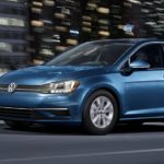 A blue 2021 Volkswagen Golf is shown driving in a city at night after leaving a Volkswagen dealership.