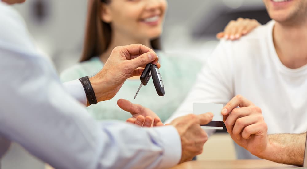 A salesman is passing a set of keys to a couple at a pre-owned Chevy dealer.