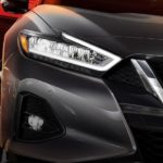 A close up of the front hood and headlights of a grey 2021 Nissan Maxima is shown on a red background.