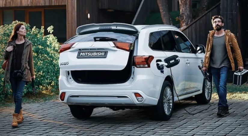 A couple is shown next to white 2022 Mitsubishi Outlander PHEV while it's charging in a driveway.