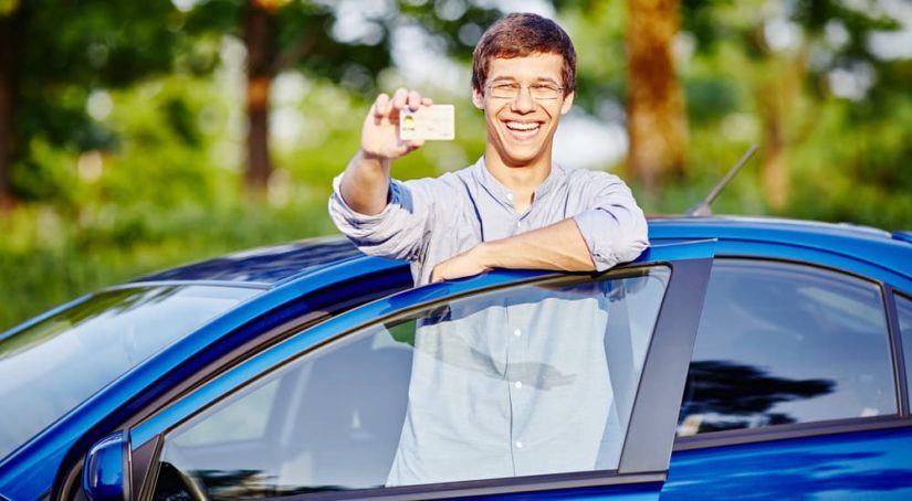 A teen driver is shown holding a license outside of a blue sedan car.