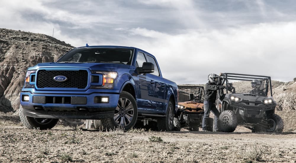 A blue 2020 Ford F-150 is shown parked near two ATVs by a remote trail.