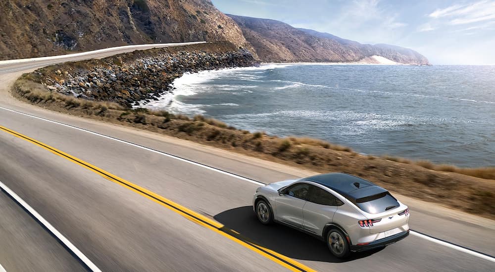 A white 2021 Ford Mustang Mach-E Premium is shown driving down a coastal road.